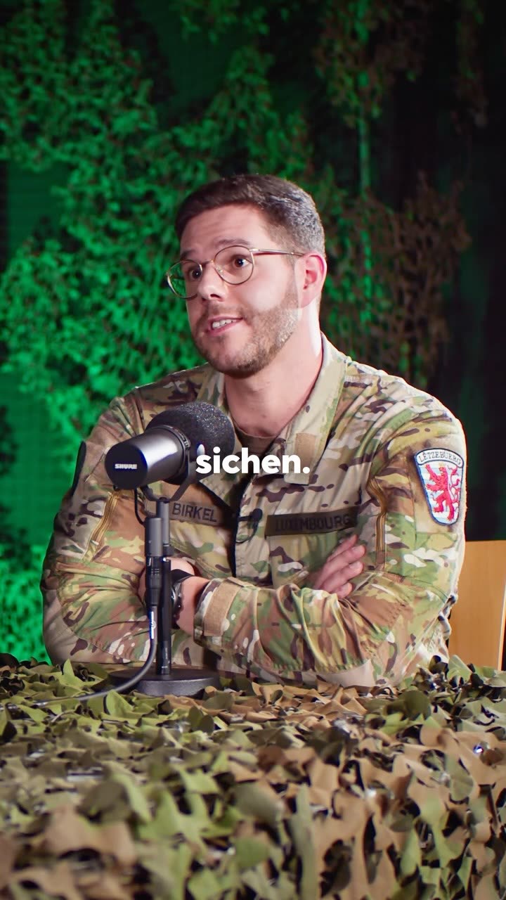 Un jeune officier en uniforme parle au micro, devant un décor camouflage